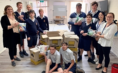 A group of teachers and children in school uniform gather around a pile of cardboard boxes, each holding zip-lock bags filled with goodies.