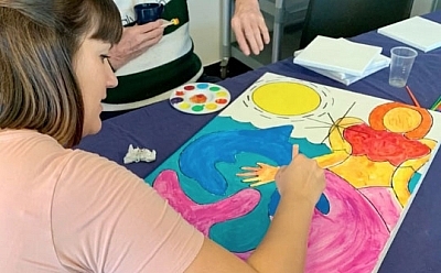 Two people sit at a table together, both focused on painting a brightly coloured picture of a mermaid and dolphin under a big sun.