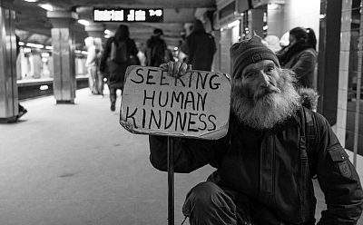 A black and white photo of a man kneeling down in a train station. He looks at the camera with gentle eyes, and holds a sign saying 'seeking human kindness'