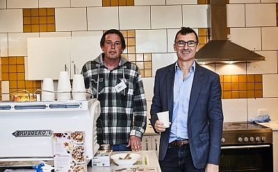 Two men stand in a kitchen behind a cafe-sized coffee machine. They both look towards the camera smiling