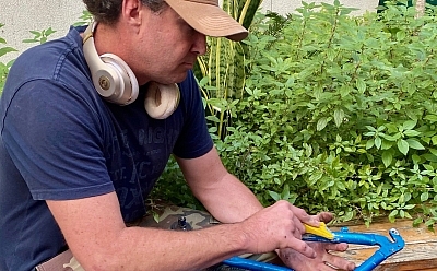 A man sits on the edge of a garden bed holding a bare bike frame and a tool. He wears headphones around his neck and a cap, and appears busy working on the bike frame