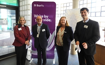 Four smartly-dressed people stand in a lobby room in front of a St Bart's sign. They all wear St Bart's name tags