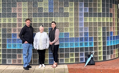 Three people stand outside a building wall made up of different coloured glass tiles. The sign above them reads 'St Bart's, we're by your side'.