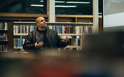 A man speaking with someone in front of a large bookcase full of books
