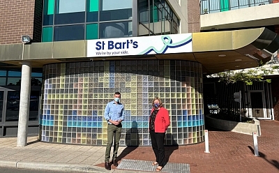 Two people stand outside a large building. The sign above them reads 'St Bart's, we're by your side'.