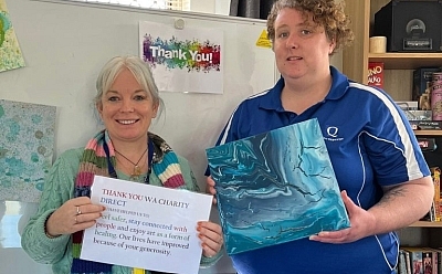 Two people stand in front of a whiteboard, one holding a sign that says ' thank you WA charity direct', and the other holding a blue artwork with wavy lines