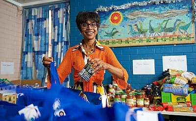 A person smiling sorting tins of food into bags. She wears happy orange clothing and stands in a bright blue room.