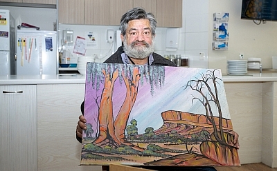 A man sits in a kitchen holding a painting of a landscape.