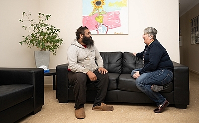 Two people sitting on a couch, facing towards each other chatting. There is a plant and a painting of a sunflower in the background.