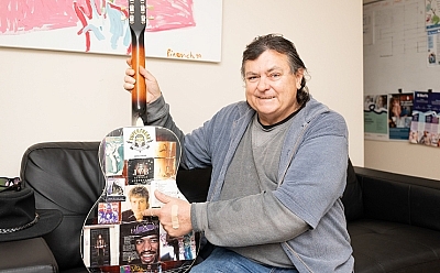A man sitting on a couch holding a guitar, pointing to the back of it which is decorated with music memorabilia.