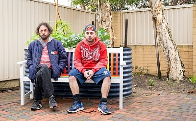 Two men sit on a colourful outdoor bench in front of a garden patch.