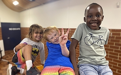 Three young kids sitting together gleefully smiling, one holds up a peace sign with their fingers